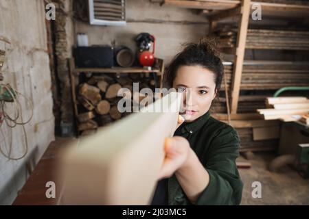 Carpentiere in bruna che controlla la tavola di legno in officina sfocata Foto Stock