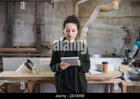 Un giovane falegname che utilizza un tablet digitale vicino alla visiera di sicurezza e un caffè per andare in officina Foto Stock