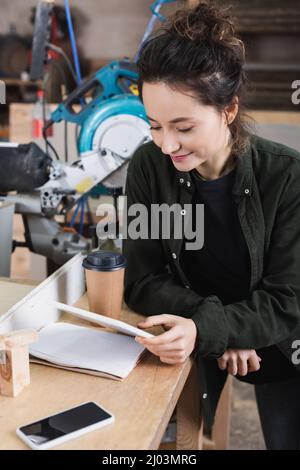 Sorridendo il lavoratore del legno utilizzando il tablet digitale vicino al cellulare e la tazza di carta in officina Foto Stock