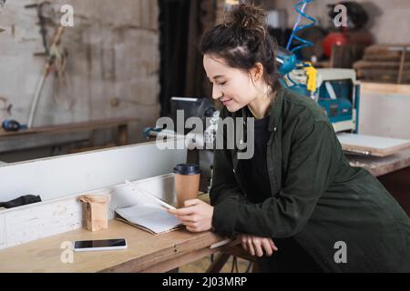 Un giovane falegname che usa un tablet digitale vicino al notebook e un caffè per andare in officina Foto Stock