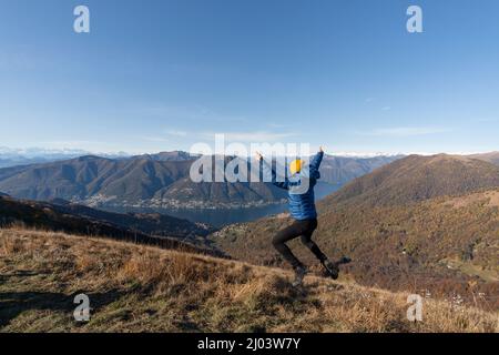 L'uomo ispanico in cima alla montagna salta con gioia ammirando il paesaggio del Lago di Como, Italia Foto Stock