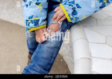 Ragazzino, che mostra il ginocchio ferito da una caduta, seduto su una panchina nel parco Foto Stock