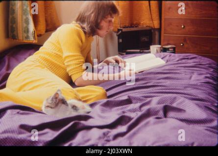Immagine archivistica, presa a metà degli anni '70 di un giovane uomo caucasico, di 23 anni, con capelli lunghi, come era la moda a quel tempo, indossando un pullover giallo e pantaloni gialli, sdraiati su un letto e leggendo un libro. Un gatto siede sul letto davanti all'uomo. Versione del modello disponibile. Foto Stock