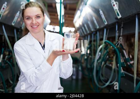 Caseificio femmina ingegnere in tunica bianca in piedi con un bicchiere di latte vicino alla linea di mungitura presso l'azienda Foto Stock