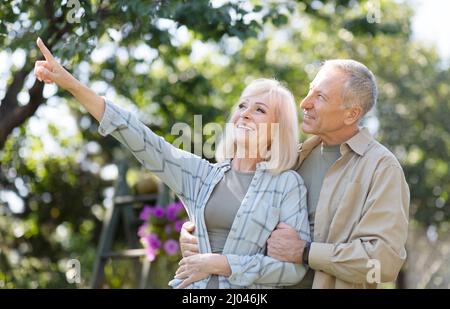 Felice coppia in pensione godendo il tempo insieme in giardino, camminando alla primavera giorno, abbracciando e donna che mostra qualcosa Foto Stock