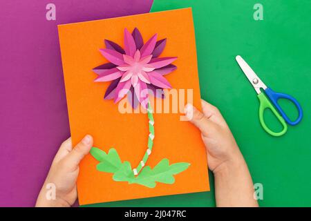 Vista dall'alto delle mani del bambino che tengono papercraft viola crysanthemum fiore fatto da bambino su foglio di lavoro arancione e forbici su sfondo viola e verde. P Foto Stock