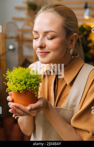 Primo piano volto di affascinante sorridente fiorista femminile in grembiule odore Soleirolia pianta che tiene in mano pentola in negozio floreale, occhi chiusi. Foto Stock