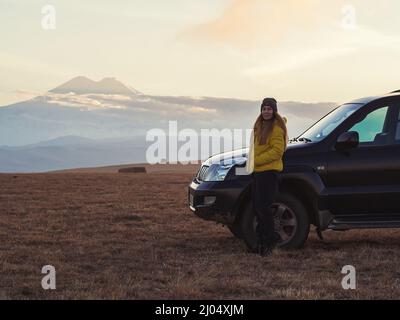 Una donna si erge vicino a un SUV in un campo di alta montagna sullo sfondo di Elbrus Foto Stock