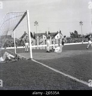 1960s, storica, rough & Tumble ......partita di calcio tra Oxford United FC e Yeovil Town FC, Manor Ground, Oxford, Inghilterra, Regno Unito. Il portiere di Yeovil in cap sittong sul terreno ferito tenendo la gamba, la grande metà centrale lo guarda, non felice, e un altro compagno di squadra nel goal che giace dalla rete con la palla. Foto Stock