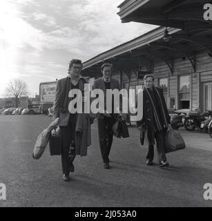 1960s, storico, di fine mandato e tre studenti universitari maschi, a piedi con i bagagli per prendere un treno a casa, Oxford, Inghilterra, Regno Unito. Due dei giovani indossano giacche sportive, una con un collo cravat, l'altra con una cravatta, mentre il terzo giovane ha un cappotto in borsello e una lunga sciarpa universitaria. Foto Stock