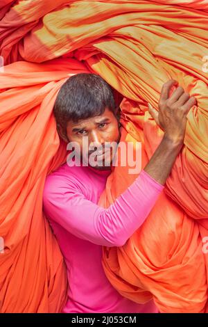 Narayanganj, Bangladesh. 16th Mar 2022. I lavoratori appendono migliaia di diversi tessuti colorati su fili di ferro legati tra un telaio di bambù e costantemente li trasformano in modo che si asciughino perfettamente in un campo allagato a Narayanganj, Bangladesh. I fili di ferro sono usati fra una struttura di bambù per generare le linee giganti di lavaggio per la parte finale del processo di tintura mentre i tessuti sono asciugati nel sole. I fili luminosi di panni blu, rosa, arancione e verde-tinti appendono sopra il campo erboso in una rete abbagliante di colori interbloccanti. Questa è la parte finale del processo di tintura dopo la quale il panno è fatto Foto Stock