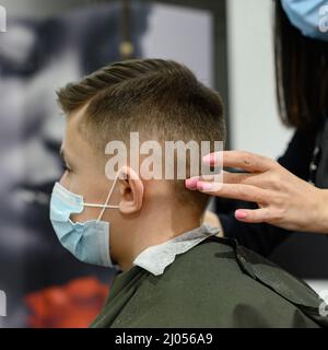 Un ragazzo adolescente ottiene un taglio di capelli in un barbiere durante una pandemia, un taglio di capelli nel salone, un cliente e un parrucchiere in maschere, un taglio di capelli del bambino con sciss Foto Stock