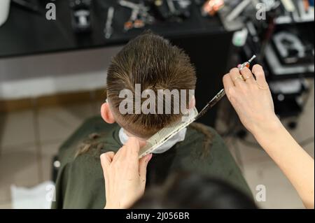 Un ragazzo adolescente ottiene un taglio di capelli in un barbiere durante una pandemia, un taglio di capelli nel salone, un cliente e un parrucchiere in maschere, un taglio di capelli del bambino con sciss Foto Stock