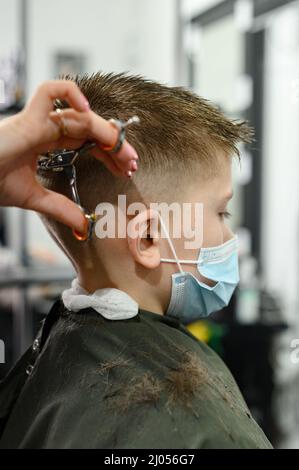 Un ragazzo adolescente ottiene un taglio di capelli in un barbiere durante una pandemia, un taglio di capelli nel salone, un cliente e un parrucchiere in maschere, un taglio di capelli del bambino con sciss Foto Stock