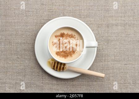 Tè piccante con latte in una tazza di porcellana bianca con cucchiaia di miele e miele su sfondo tessile Foto Stock