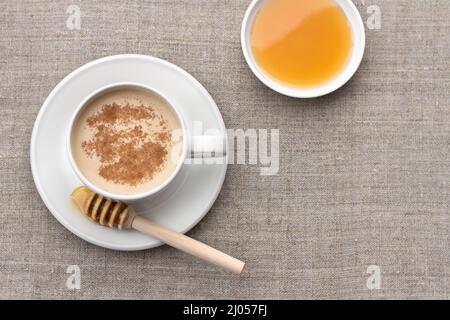Tè piccante con latte in una tazza di porcellana bianca con cucchiaia di miele e miele su sfondo tessile Foto Stock