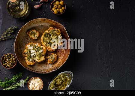 Bistecca di cavolfiore con spezie, salsa di chimichurri, fiocchi di mandorle. Capperi, olive, erbe, fianco a fianco. Sfondo scuro. Cibo vegetariano. CopySpace. Foto Stock