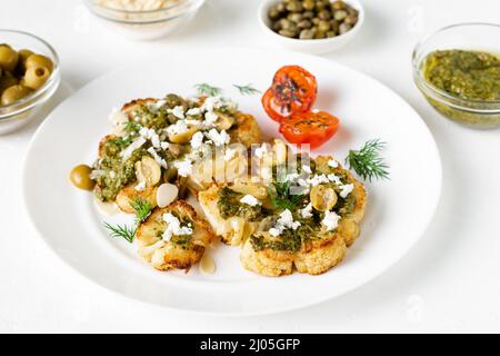 Bistecca di cavolfiore con spezie, salsa di chimichurri, fiocchi di mandorle, olive, pomodori ciliegini fritti e capperi su un piatto bianco. Cibo vegetariano. Foto Stock