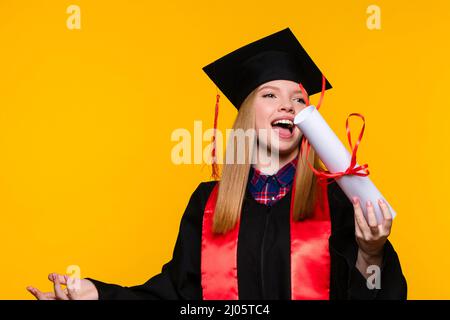 Laureato con cappello di laurea e diploma su sfondo giallo. La giovane donna bionda che indossa il cappuccio di graduazione e l'abito da cerimonia lancia certificato legato con nastro rosso. Concetto di istruzione Foto Stock