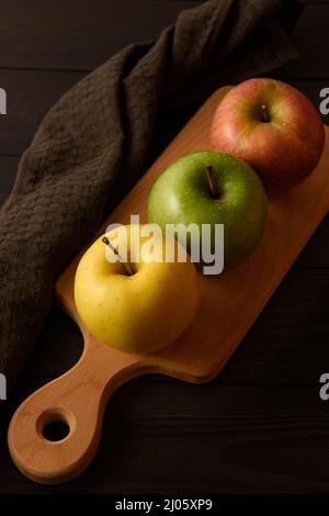 Le mele gialle, verdi e rosa si trovano su un asse di legno accanto ad un tovagliolo di cotone marrone su uno sfondo di legno marrone. Foto Stock