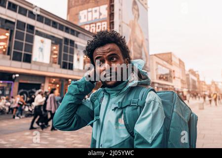 Ragazzo con consegna curly con giacca di cappotto turchese e zaino da cucina in strada a Glasgow, Scozia Foto Stock