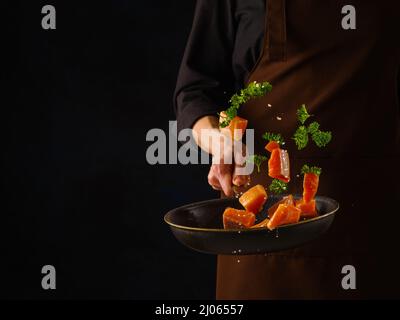 Uno chef professionista in uniforme scura prepara pezzi di trota rossa, salmone con verdure ed erbe in una padella su sfondo nero. Levit Foto Stock