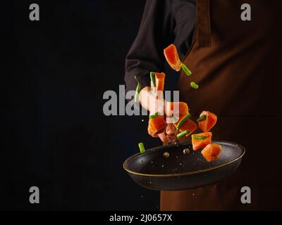 Uno chef professionista prepara pezzi di trota, salmone con verdure. Cucina di pesce, cibo vegetariano sano su sfondo nero in volo congelato. B Foto Stock