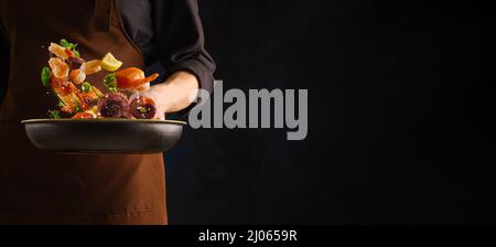 Uno chef professionista in uniforme scura prepara frutti di mare assortiti - pezzi di pesce rosso, polpo, gamberi in una padella su sfondo nero. Levitazione. Mare Foto Stock