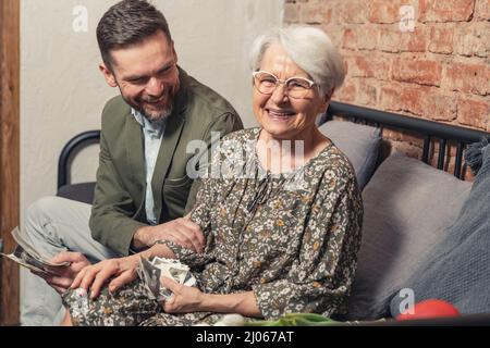 felice sorridente ridente nonna dai capelli corti siede su un divano e guarda le vecchie immagini con il suo figlio imprenditore adulto. Foto di alta qualità Foto Stock