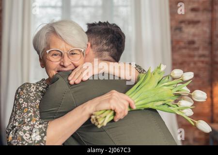 allegro sorridente anziano pensionato donna abbracciando il suo figlio uomo d'affari adulto e tenendo bouquet di tulipani bianco medio colpo. Foto di alta qualità Foto Stock