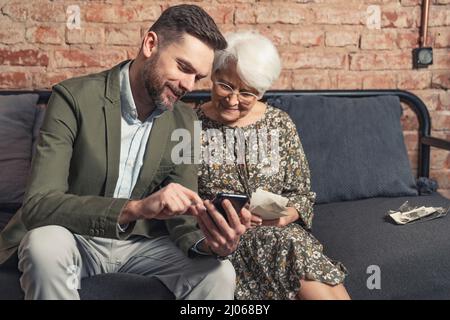 donna pensionato anziana dai capelli grigi seduta su un divano e guardando lo smartphone del figlio adulto millenario. Foto di alta qualità Foto Stock