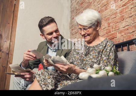 Vivace vecchia donna grigio-capelli pensa volentieri al passato e mostra il suo nipote adulto vecchie fotografie il giorno del nonno. Foto di alta qualità Foto Stock