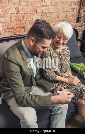colpo verticale di un uomo di mezza età in tuta e la sua anziana madre dai capelli grigi seduta su un divano e parlando di eventi passati. Foto di alta qualità Foto Stock