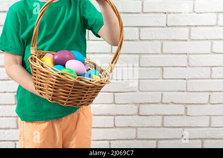 Ragazzo senza volto con uova di Pasqua colorate in cestino di vimini vicino a muro di mattoni bianchi Foto Stock