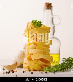 Su sfondo bianco, pasta italiana in un interessante servizio verticale, aglio, rametti di rosmarino e una bottiglia di olio d'oliva. Ricette di cucina italiana, f Foto Stock