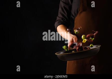 Uno chef professionista in un'uniforme scura cuoce il polpo con le verdure in una padella su sfondo nero. Ricette di frutti di mare. Cucina piatti di pesce. Esotica Foto Stock