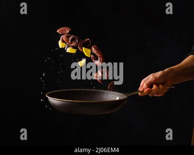 Uno chef professionista prepara polpo con fette di limone in una padella su sfondo nero. Levitazione. Ricette orientali e asiatiche di pesce, ristorante Foto Stock