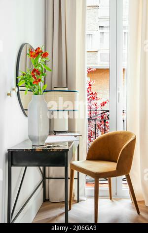 Scrivania in acciaio con piano in legno, grande vaso grigio con fiori, specchio circolare e sedia rivestita in velluto arancione e balcone con tende e rete Foto Stock