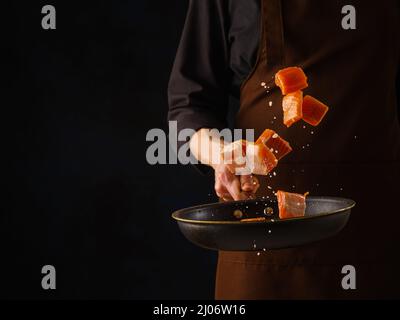 Uno chef professionista in uniforme nera prepara pezzi di pesce rosso - salmone, trota in una padella. Levitazione. Sfondo nero. Vegetariano sano f Foto Stock