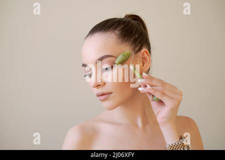 Primo piano ritratto caucasico bella donna facendo ascensore sollevamento levigare massaggio anti-invecchiamento con rullo di pietra verde, isolato su sfondo beige con spazio copia. Concetto di cura della pelle del viso Foto Stock