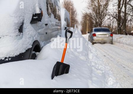 Pala da neve in plastica di fronte al minivan coperto di neve al sole della mattina d'inverno e auto d'argento che passa in background sfocato Foto Stock