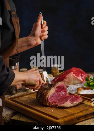 Lo chef prepara carne, manzo. Le mani dello chef preparano carne alla bistecca o prosciutto e aggiungono sale e pepe sullo sfondo scuro per il testo del menu del ristorante o del reci Foto Stock