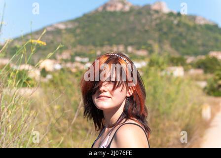 Giovane donna con capelli neri arancioni guardando la macchina fotografica mentre si trova di fronte a una montagna vicino cala millor, maiorca, spagna Foto Stock