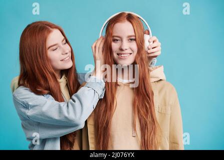 Vita ritratto di bella donna cacuasiana con capelli rossi mettendo cuffie bianche sulla testa delle sue sorelle gemelle, sfondo blu Foto Stock