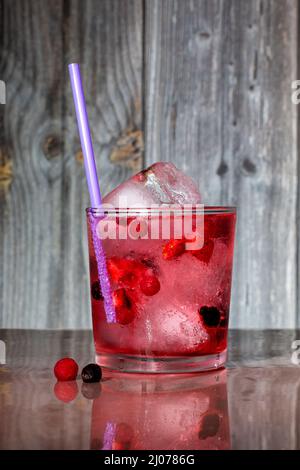 Un bicchiere cilindrico con una combinazione di gin, tonico, ghiaccio e frutti rossi con una cannuccia. E' collocato su un tavolo marrone dove si riflette e sul retro Foto Stock