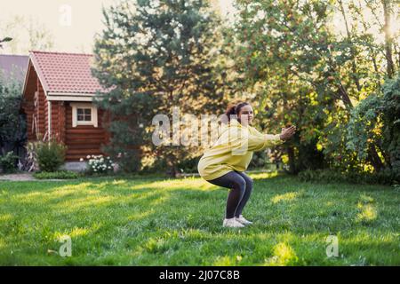 Donna su erba verde facendo yoga squatting in abiti sportivi guardando la macchina fotografica sul cortile pieno di luce del sole con casa di campagna e alti alberi in Foto Stock