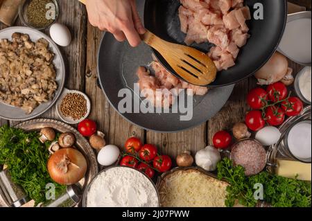 Lo chef versa a mano il filetto di pollo tritato in una padella per preparare il ripieno su un tavolo di legno con una varietà di ingredienti di fondo. Concetto di cottura proc Foto Stock