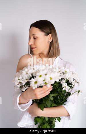 Giovane bella carina dolce bella sorridente donna con tenere un bouquet di fiori freschi bianchi su sfondo bianco parete Foto Stock