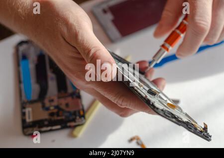 Repair of smartphones concept. A man unscrews the screws fixing the display from the board. Part of a series. Inside the room. Selective focus. Foto Stock