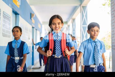 Bambino felice ragazza in uniforme in piedi al corridoio della scuola mentre altri bambini passano - concetto di fiducia, istruzione, crescita infantile e sviluppo. Foto Stock